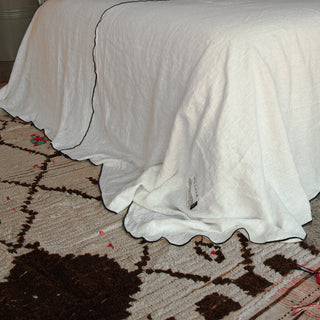 detalle de colcha blanca de lino con vivo de color negro sobre alfombra de lana natural.