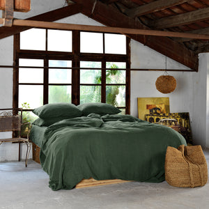 Cama vestida con juego de lino verde oscuro. Sobre gran ventanal de madera con techo de vigas antiguo.