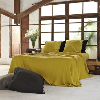 Cama vestida de lino mostaza con almohadones negros. Cama en espacio amplio de madera y grandes ventanales.